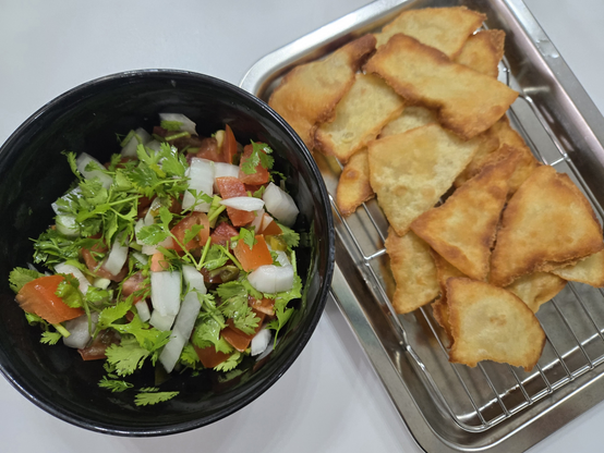 검정 그릇에 고수, 토마토, 양파가 주로 되어있는 살사가 담겨있고, 금속 밧드위에 적당한 크기로 적당한 크기로 잘라서 노릇하게 팬프라잉된 또르띠야가 있다.